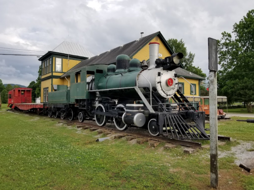 A vintage steam locomotive displayed next to a yellow train station building on grassy grounds.