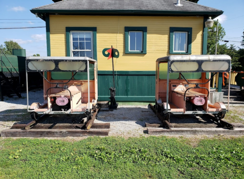 Two vintage rail cars parked beside a yellow building, with green trim and a clear blue sky in the background.