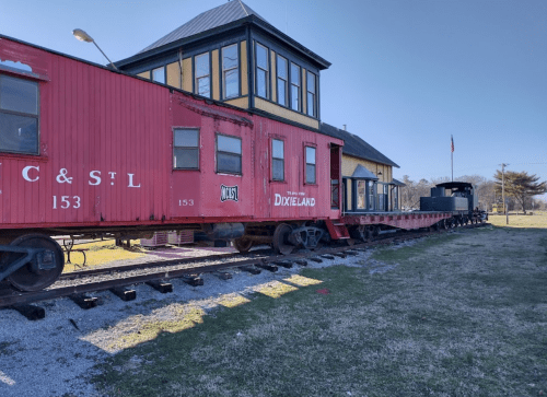 A red train car and a black locomotive on tracks near a historic building, set in a grassy area.