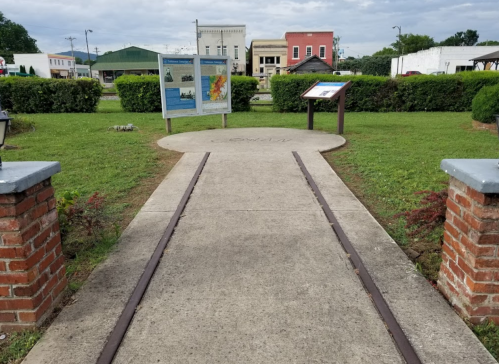 A pathway with brick edges leads to informational signs in a grassy area, with buildings visible in the background.