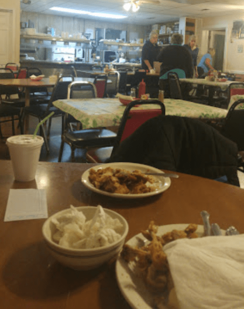 A cozy diner interior with tables, a kitchen in the background, and plates of food on a wooden table.