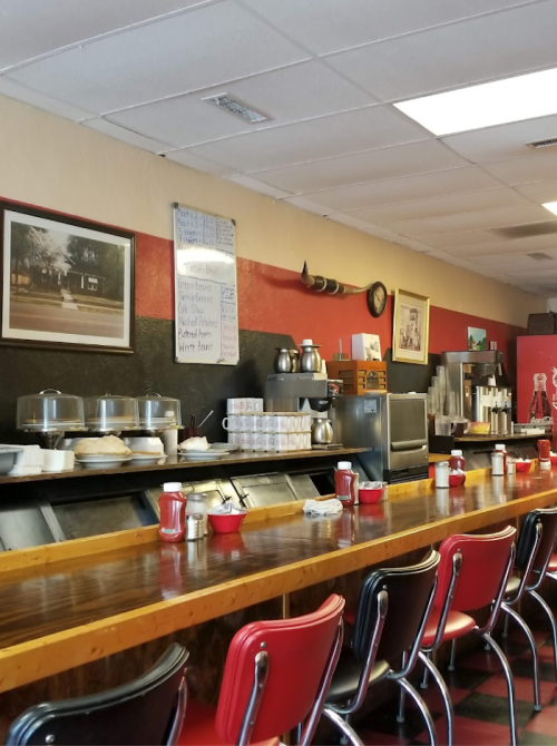 A cozy diner interior with a wooden counter, red chairs, and vintage decor, featuring food displays and coffee pots.