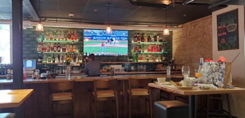 A bar scene with a bartender, shelves of liquor, and a TV showing a baseball game. Tables are set with drinks and food.