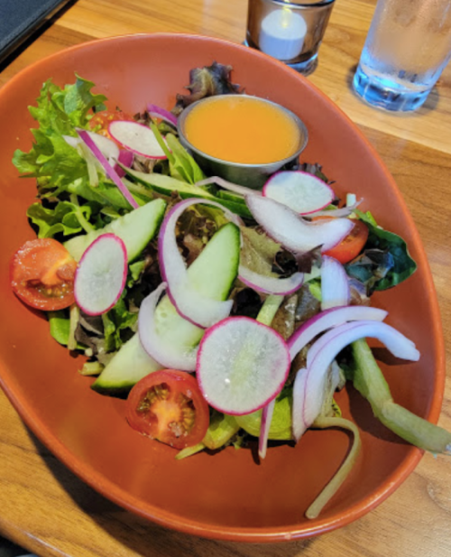 A colorful salad with mixed greens, sliced cucumbers, tomatoes, radishes, and a small bowl of dressing.