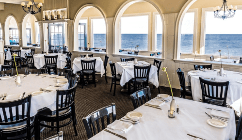 A bright restaurant with white tablecloths and black chairs, featuring large windows overlooking the ocean.