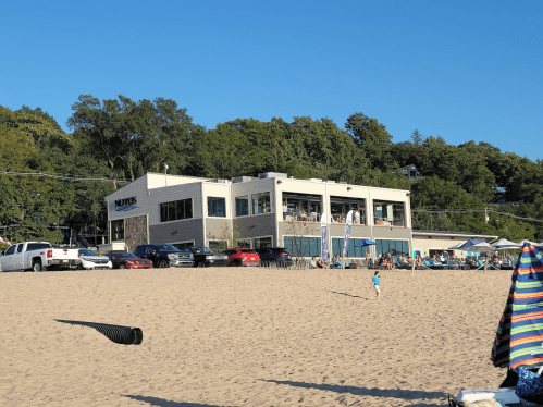 A beach scene featuring a building with large windows, surrounded by trees, and cars parked on the sandy shore.