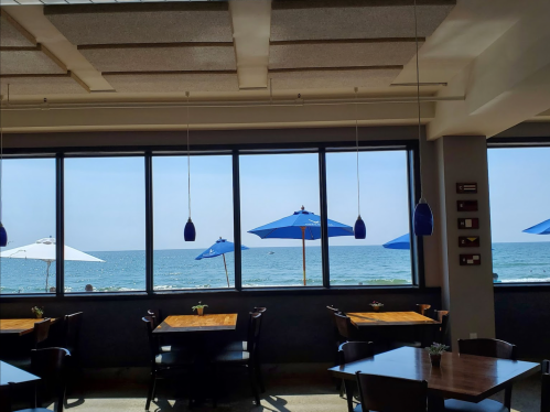 A seaside restaurant with wooden tables and blue umbrellas visible through large windows overlooking the ocean.