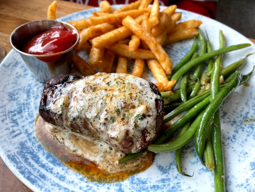 A plate with a grilled steak topped with garlic butter, served with fries, green beans, and a side of ketchup.