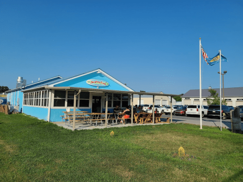 A blue restaurant with outdoor seating, flags flying, and cars parked nearby on a sunny day.