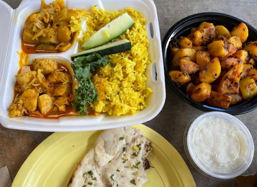 A takeout meal featuring yellow rice, chicken with vegetables, roasted potatoes, naan bread, and a small container of sauce.