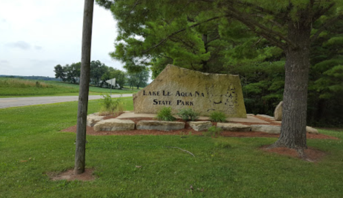 Sign for Lake Le Aqua Na State Park, featuring a large stone structure surrounded by trees and grass.