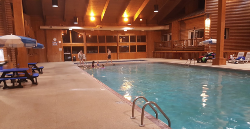 Indoor swimming pool with wooden beams, lounge chairs, and children playing in the water. Soft lighting creates a warm atmosphere.