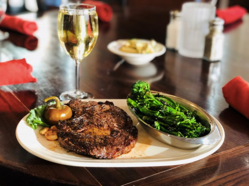 A plated steak with broccolini and a glass of white wine on a wooden table, with red napkins in the background.