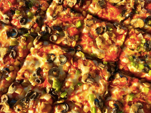 A close-up of a pizza topped with cheese, olives, mushrooms, green peppers, and diced onions, cut into squares.