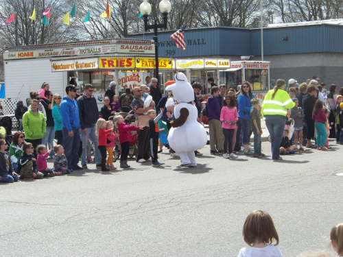 A crowd gathers at a parade, watching a costumed character in a white outfit dance among children and families.