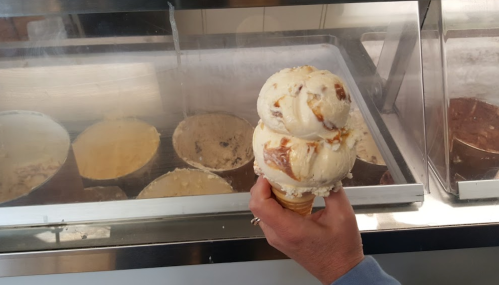 A hand holding a double scoop of ice cream in a cone, with various ice cream tubs in the background.