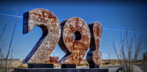 A large, rust-colored sculpture of the number "29!" against a clear blue sky in a desert landscape.
