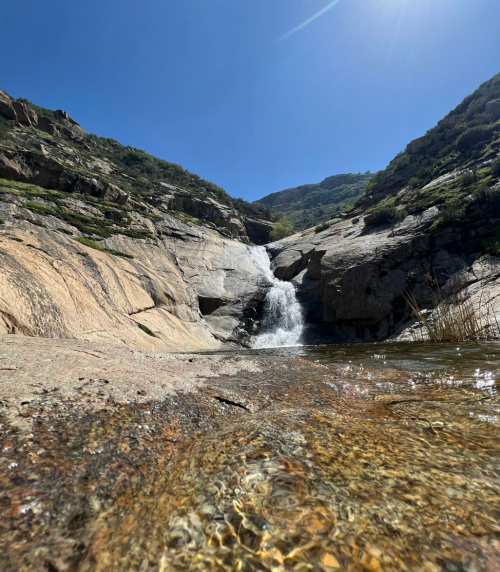 A serene waterfall cascading over rocky terrain, surrounded by mountains under a clear blue sky.