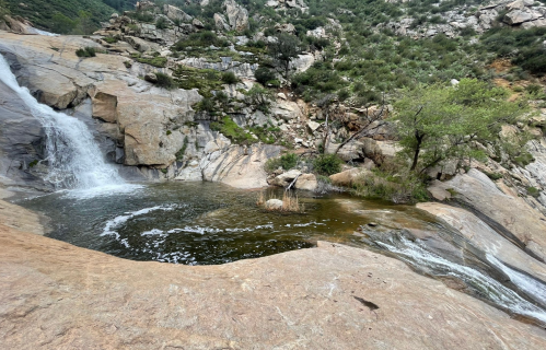 A serene waterfall cascades over rocky terrain into a calm pool, surrounded by lush greenery and rugged hills.