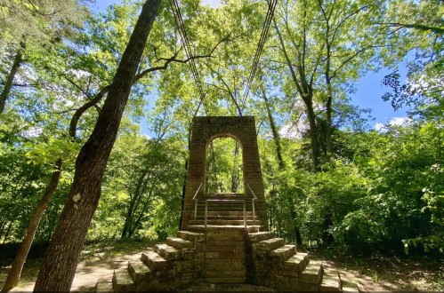 A stone archway with steps surrounded by lush green trees and a bright blue sky.