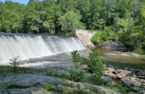 A serene view of a waterfall cascading over a dam, surrounded by lush green trees and rocky riverbanks.