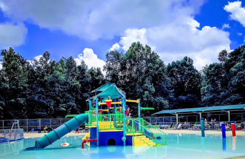Colorful water play structure with slides in a pool, surrounded by trees and blue sky with fluffy clouds.