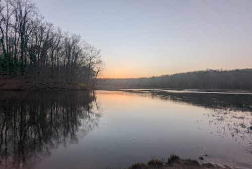 Serene lake at dusk, reflecting trees and a soft sunset glow, with calm waters and a peaceful atmosphere.