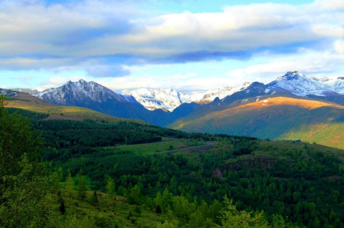 A scenic view of mountains with snow-capped peaks, lush green valleys, and a partly cloudy sky.