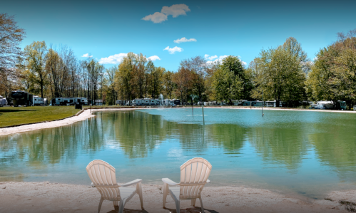 Two white Adirondack chairs face a serene lake surrounded by trees and RVs under a clear blue sky.