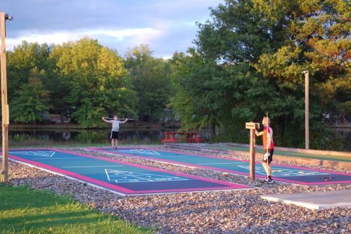 Two people playing shuffleboard on an outdoor court by a lake, surrounded by trees and a picnic area.