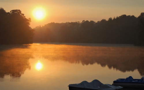 Sunrise over a misty lake, with golden reflections and silhouettes of trees in the background. Paddle boats in the foreground.