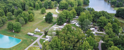 Aerial view of a campground with RVs, green trees, and a small pond surrounded by grassy areas.