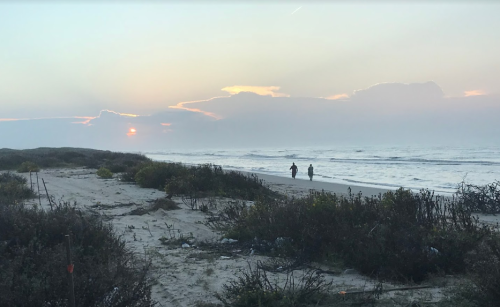 Two people walk along a sandy beach at sunset, with gentle waves and a cloudy sky in the background.