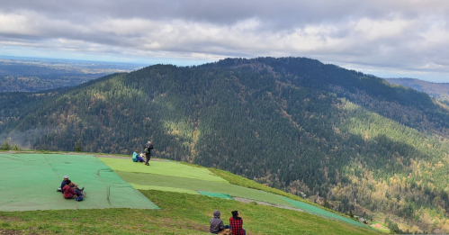 A scenic view from a mountain top, with people relaxing on green patches overlooking a lush, forested landscape.