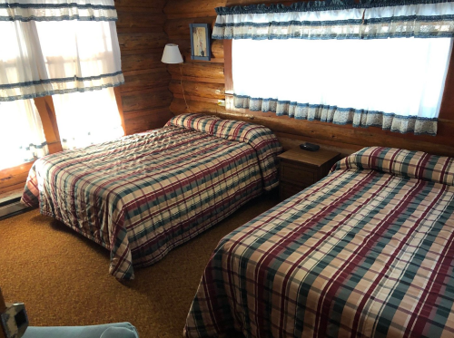 Cozy cabin bedroom with two plaid-covered beds, wooden walls, and large windows letting in natural light.