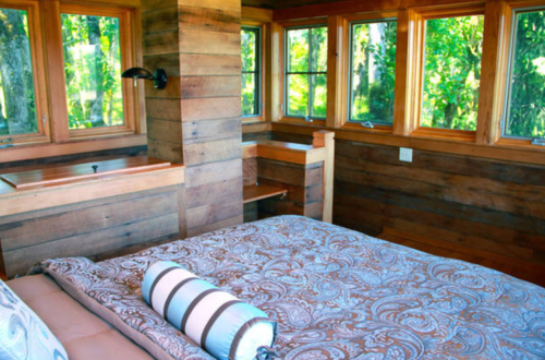 Cozy wooden bedroom with large windows, a patterned bedspread, and a decorative pillow. Natural light fills the space.