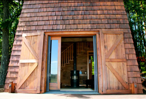 A wooden cabin with a triangular roof and open double doors, revealing a cozy interior and a staircase.