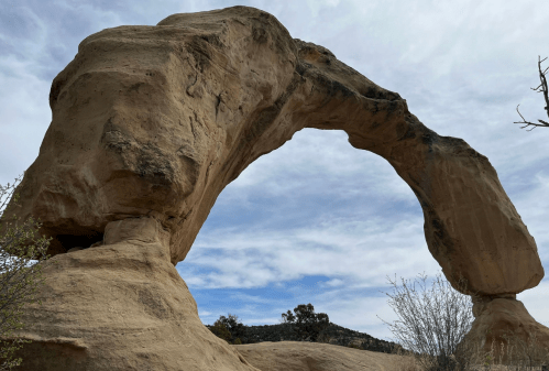Natural rock arch against a cloudy sky, surrounded by sparse vegetation and distant hills.