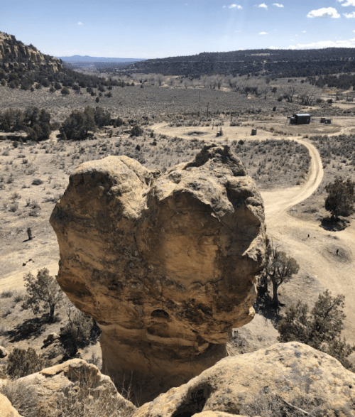 A large rock formation stands prominently in a desert landscape, with sparse vegetation and a winding dirt road nearby.