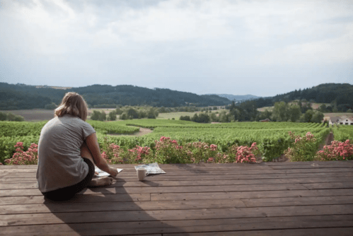 A person sits on a wooden deck, painting, with a scenic view of vineyards and hills in the background.