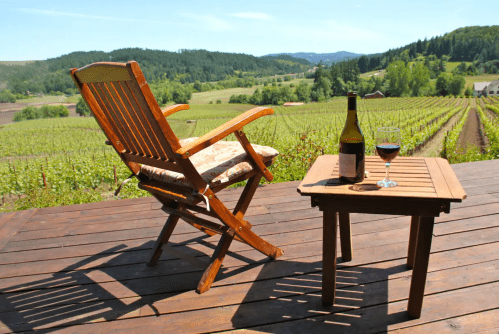 A wooden chair and table on a deck overlooking a vineyard, with a bottle of wine and a glass.