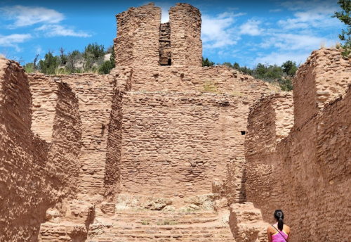 A person stands before ancient stone ruins under a blue sky, surrounded by trees and rocky terrain.