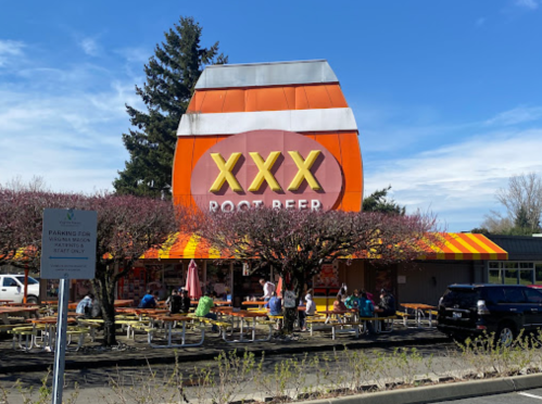 A colorful restaurant with a large root beer barrel sign, outdoor seating, and trees in bloom.