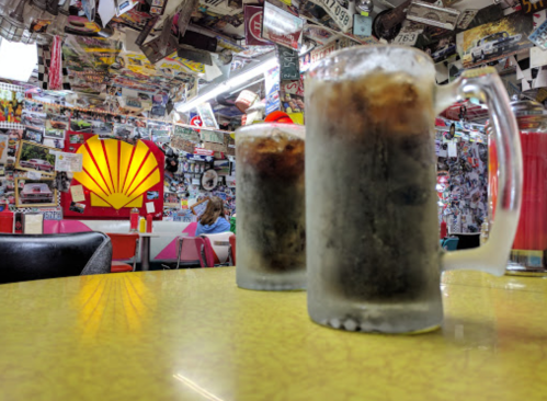 Two frosty mugs of soda sit on a yellow table, with a colorful, vintage-themed diner in the background.