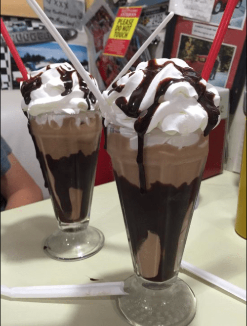 Two tall glasses of chocolate milkshakes topped with whipped cream and chocolate syrup, served with straws.