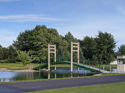 A green pedestrian bridge spans a calm pond, surrounded by lush trees and a clear blue sky.