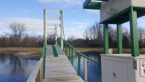 A wooden suspension bridge leads over a calm body of water, with trees in the background and a small structure nearby.