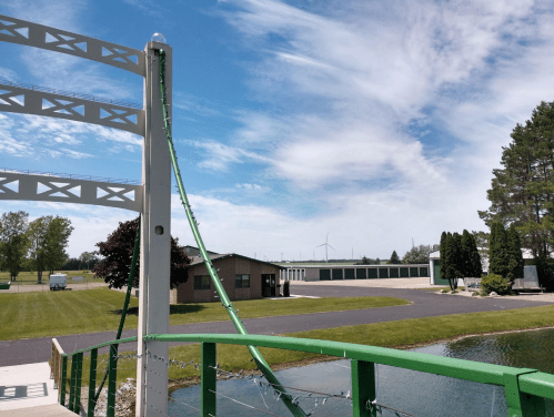 A green bridge with a curved support, overlooking a pond and a storage facility under a blue sky with scattered clouds.