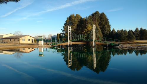 A green suspension bridge reflects in calm water, surrounded by trees and a building under a clear blue sky.