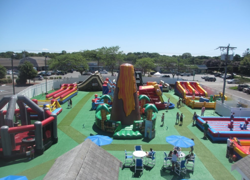 A vibrant outdoor inflatable park featuring a volcano slide, bounce houses, and people enjoying the attractions.